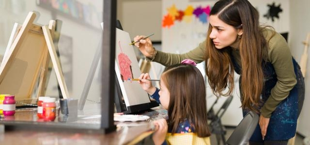 attractive hispanic teacher is teaching pretty child girl how use paint brush canvas art class scaled