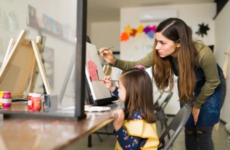 attractive hispanic teacher is teaching pretty child girl how use paint brush canvas art class scaled